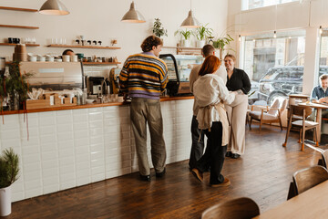 A group of adults stands and talks at the counter of a modern cafe. A woman in an apron speaks with customers. Another person sits at a window table.