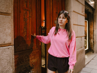 Fototapeta premium A young woman dressed in a bright pink sweatshirt and black shorts stands outdoors as she opens a large wooden door on a city street lined with stone walls and urban features.