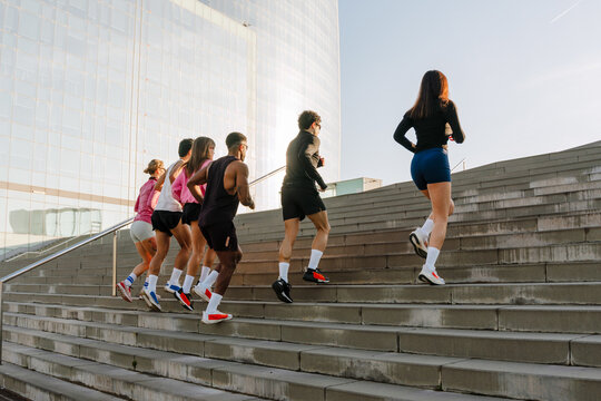 A multinational group of young people running up concrete stairs outside a modern building on a sunny day, wearing athletic clothing, exercising together as part of an outdoor training session.