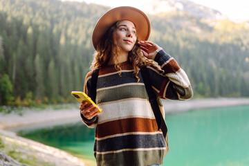 Young woman with phone and hat sitting near mountain lake at sunrise. Cute female traveler with...