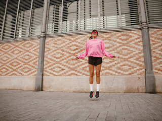 A white adult woman wearing a pink sweatshirt and black shorts is jumping rope outdoors in front of...