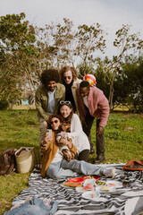 A group of multiracial friends, including Asian, Black, and White men and women, sits and stands together on a picnic blanket outdoors in a park, taking selfies with a dog and eating lunch.