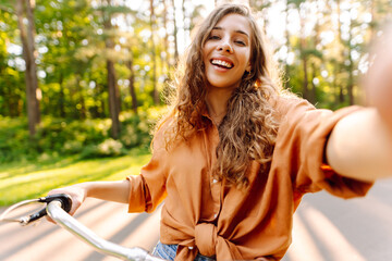 A beautiful woman takes a selfie on a bicycle in a sunny park. A young woman blogs and enjoys a...