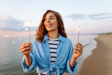 Happy woman holding sparklers on the beach. Young woman having fun holding sparklers at sunset,...