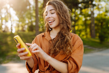 Portrait of cheerful woman with smartphone texting and blogging in sunny park. Young woman relaxing outdoors and texting with phone in hand. Blogging and technology concept.