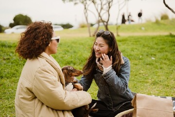Two multiracial adult women sit on the grass in a park, smiling and talking while holding a small...