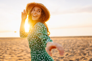 Portrait of happy woman in straw hat walking on sunset beach. Young woman in dress enjoying sea...