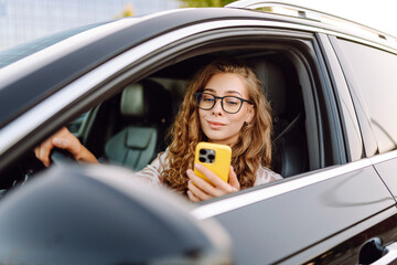 Portrait of a young female driver driving a car with a phone in her hands. Beautiful woman with a...