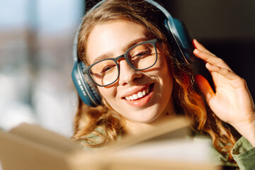 Close-up of a young woman with wireless headphones reading a book in a cozy room. Happy woman...