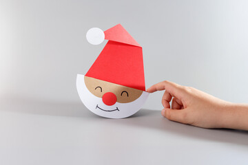 Handmade paper Santa Claus face with red hat and white beard touched by child finger. Christmas kids craft made from colored paper on grey background. Winter holiday and DIY concept. copy space,