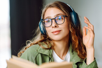 Close-up of a young woman with wireless headphones reading a book in a cozy room. Happy woman...