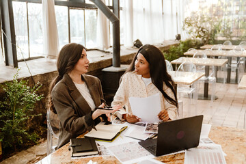 The young White and Black women sit at a table in a modern cafe, discussing documents with a laptop...
