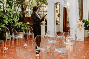 A woman stands in a beautiful indoor space filled with plants, transparent chairs, and elegant decor, taking notes with a planchette. The concept of an intimate event setup.