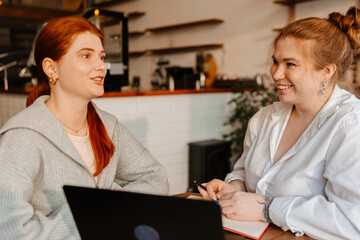 A young White female cafe owner smiling while discussing work details with a young White female worker sitting together at a table inside a modern cafe during the daytime, using a laptop and notebook.