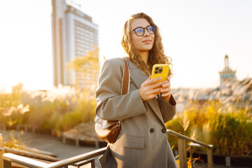Portrait of young woman in glasses and with phone enjoying sunset with city view. Smiling woman...