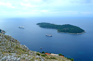 Cruise ship and sailing vessel anchored near forested islands in calm blue sea, viewed from rocky...