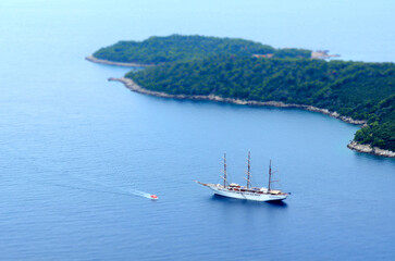 Cruise ship and sailing vessel anchored near forested islands in calm blue sea, viewed from rocky coastal cliff