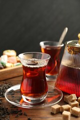 Traditional Turkish tea in glass cups, teapot, dry leaves, brown sugar and sweets on wooden table, closeup