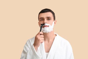 Handsome young man with razor shaving his face on beige background