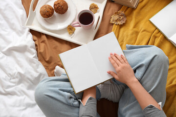 Woman reading book with tray and autumn leaves on bed, top view