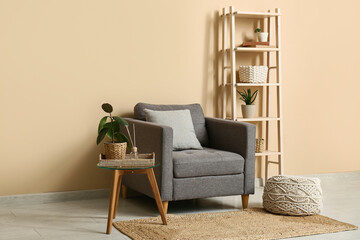 Stylish grey armchair with soft pouf and table near beige wall in room