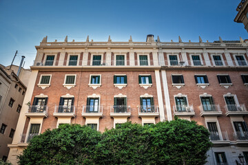 Brick-and-stucco apartment façade in Ciutat Vella displays green-shuttered windows, iron balconies, neo-classical cornice above dense shrubbery, illustrating city-centre housing real estate heritage.