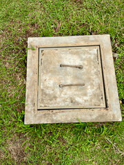 Grey cement control tank cover with iron handle on top in a garden with green grass around it, drainage control tank concept