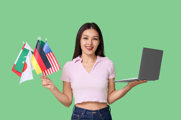 Young Asian woman with flags of different countries and laptop on green background