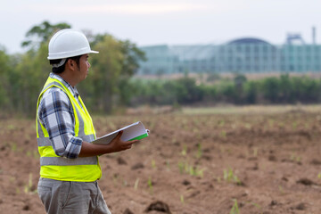 A factory worker studies a project document while standing in a rural farming area with an industrial factory in the background. Concept of development, industry meets agriculture.