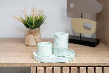 Two elegant concrete candle holders with a soft green hue are arranged on a round tray on a wooden table. A small plant in a tied fabric pot adds a touch of nature to the setting.