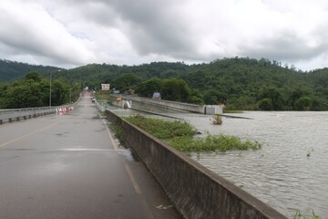 Fototapeta premium Flooded Bridge Over River in Thailand: Heavy Rainfall Causes Overflow, Natural Disaster, Green Mountains Landscape