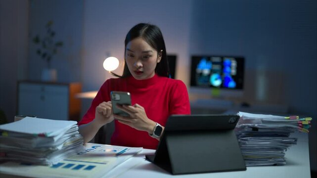 A woman is sitting at a desk with a pile of papers and a laptop. She is looking at her cell phone