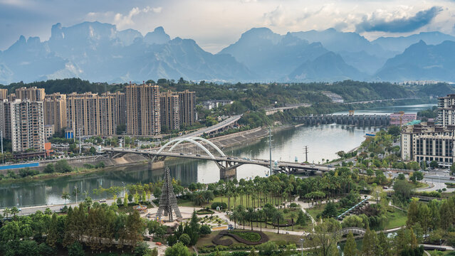 Urban landscape. View from the height. A calm river. Cars are driving over the bridge. The park area has green lawns, palm trees, a playground with carousels. Multi-storey buildings. Mountains