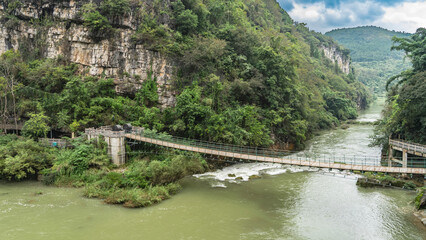 A pedestrian suspension bridge spans over a mountain river. Water foams on boulders and rapids in...