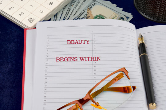 A red notebook opened to a motivational quote about beauty, placed on a desk covered with glasses and several banknotes, creating an inspiring work environment