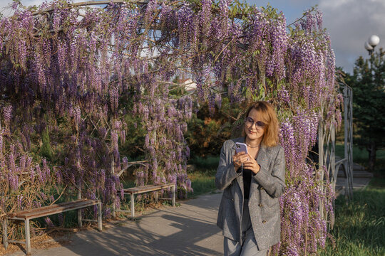 Wisteria, Woman, Phone - Woman in a gray jacket using her phone while walking in a park with a large wisteria arbor. - Powered by Adobe