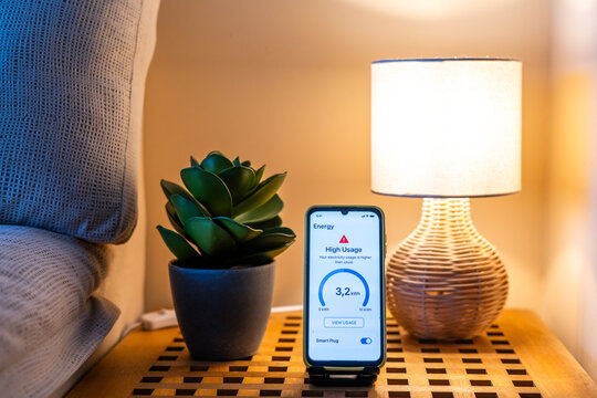 Home energy consumption monitoring app displaying high usage alert on smartphone placed on nightstand next to a lit lamp and a plant, promoting energy saving awareness and smart home technology