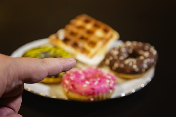A finger pointing at a plate with sweets — doughnuts and Belgian waffles — on a black background.