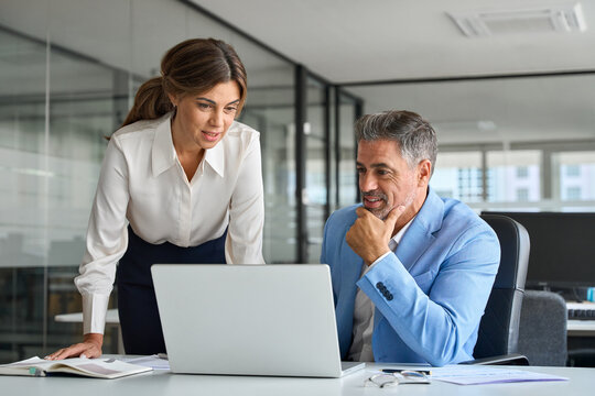 Professional mature business man and woman leaders colleagues talking working on computer at work desk. Two busy professional executives company team working in corporate office using pc laptop.