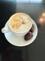 A white cup of frothy Arabic coffee served with two dates on a white saucer placed on a black...
