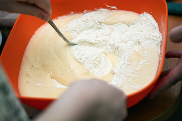 Mixing flour with liquid egg-milk mixture using a metal spoon to prepare homemade dough in a...