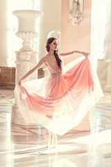 Beautiful girl with long curly hair in transparent pink dress. Ballerina on pointe dancing in sunlight from windows