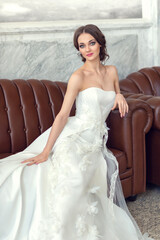 Portrait of a beautiful girl-bride in a wedding dress. A slender young woman with bare shoulders sits on the sofa. Wedding makeup and hairstyle. A beautiful face with big green eyes.