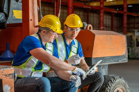Two workers in hard hats and safety vests are sitting on a forklift, looking at a tablet.