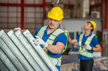 A man in a yellow hard hat and blue vest is inspecting metal sheets in a warehouse.