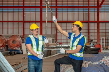 Two workers wearing hard hats and safety vests are working together to lift a heavy load with a chain hoist.