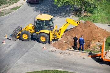 A yellow excavator dug up a road to lay electrical cables, workers stand near the hole, industry. Copy space for text