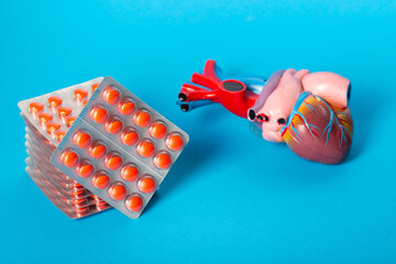 Stack of red pills against the background of a heart model and a blue background. Concept of treating diseases in cardiology, angina. Vitamins for the heart. Pills for arrhythmia. Copy space for text
