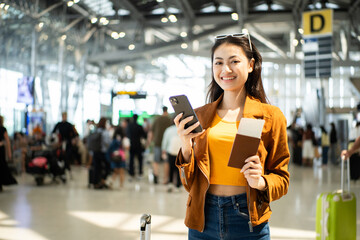 happy young asian woman traveler holding boarding pass ticket, passport and smartphone at the airport terminal with her luggage and smiling at camera, cheerful tourist female having holiday trip.