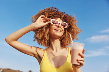 Happy woman with curly hair wearing sunglasses and holding a milkshake against a blue sky